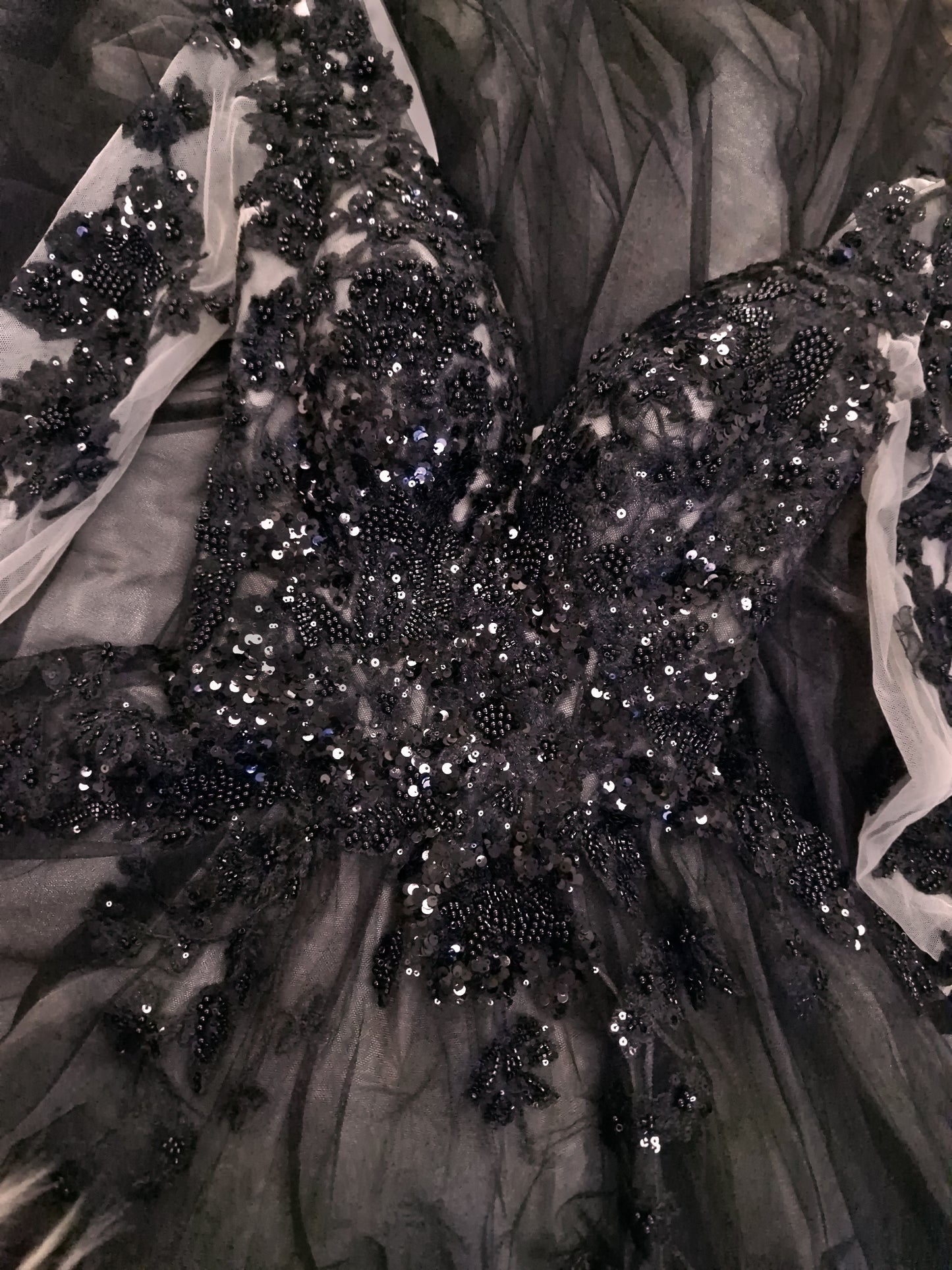 Close-up of a black tulle ball gown with intricate beading, sequins, and lace appliques, showing the detailed texture of the bodice and sleeves.