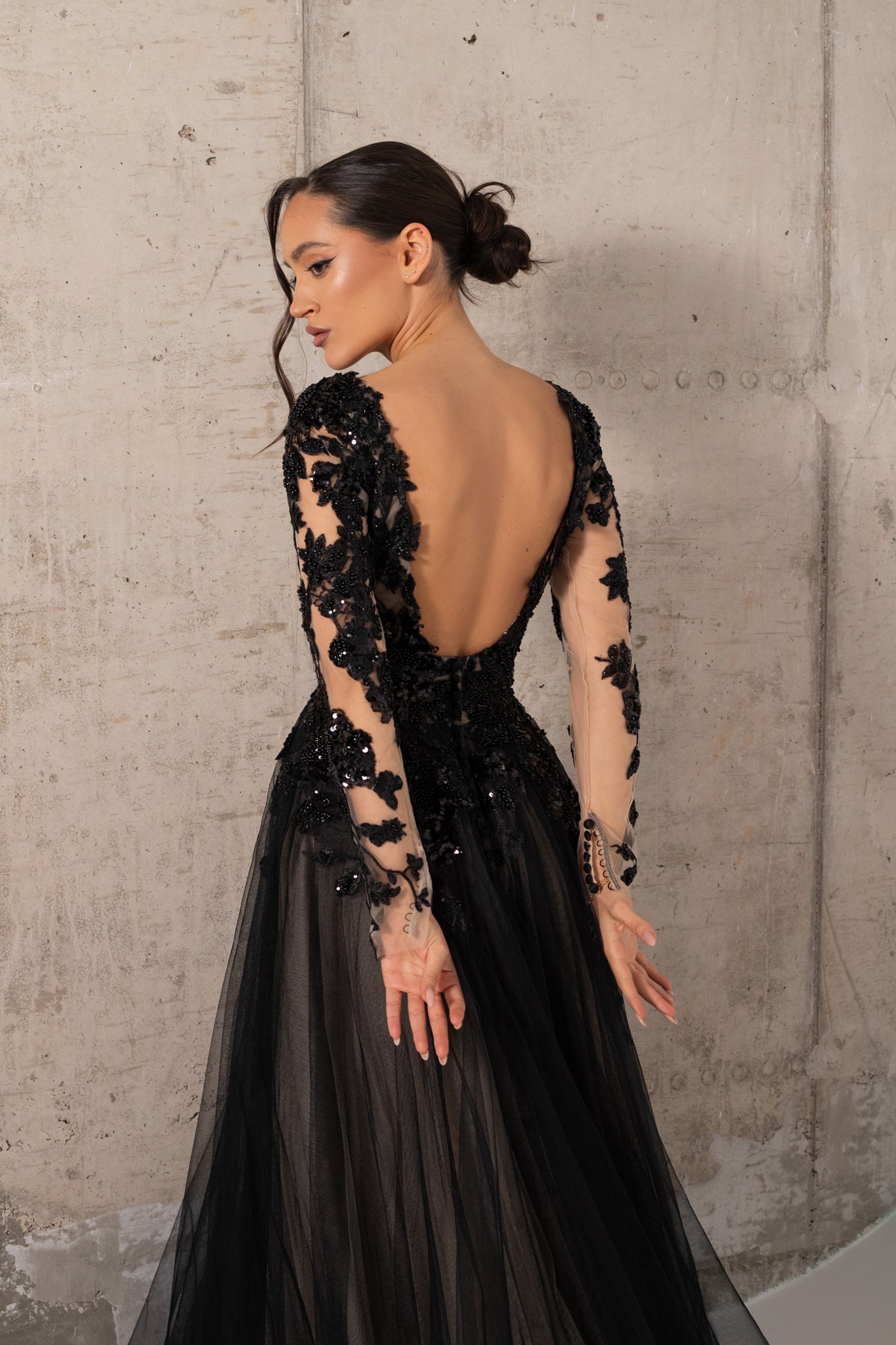 A woman modeling a black evening gown from the back, featuring long sheer sleeves with black floral lace applique, a low open back, and a flowing black tulle skirt, posed against a textured concrete wall.