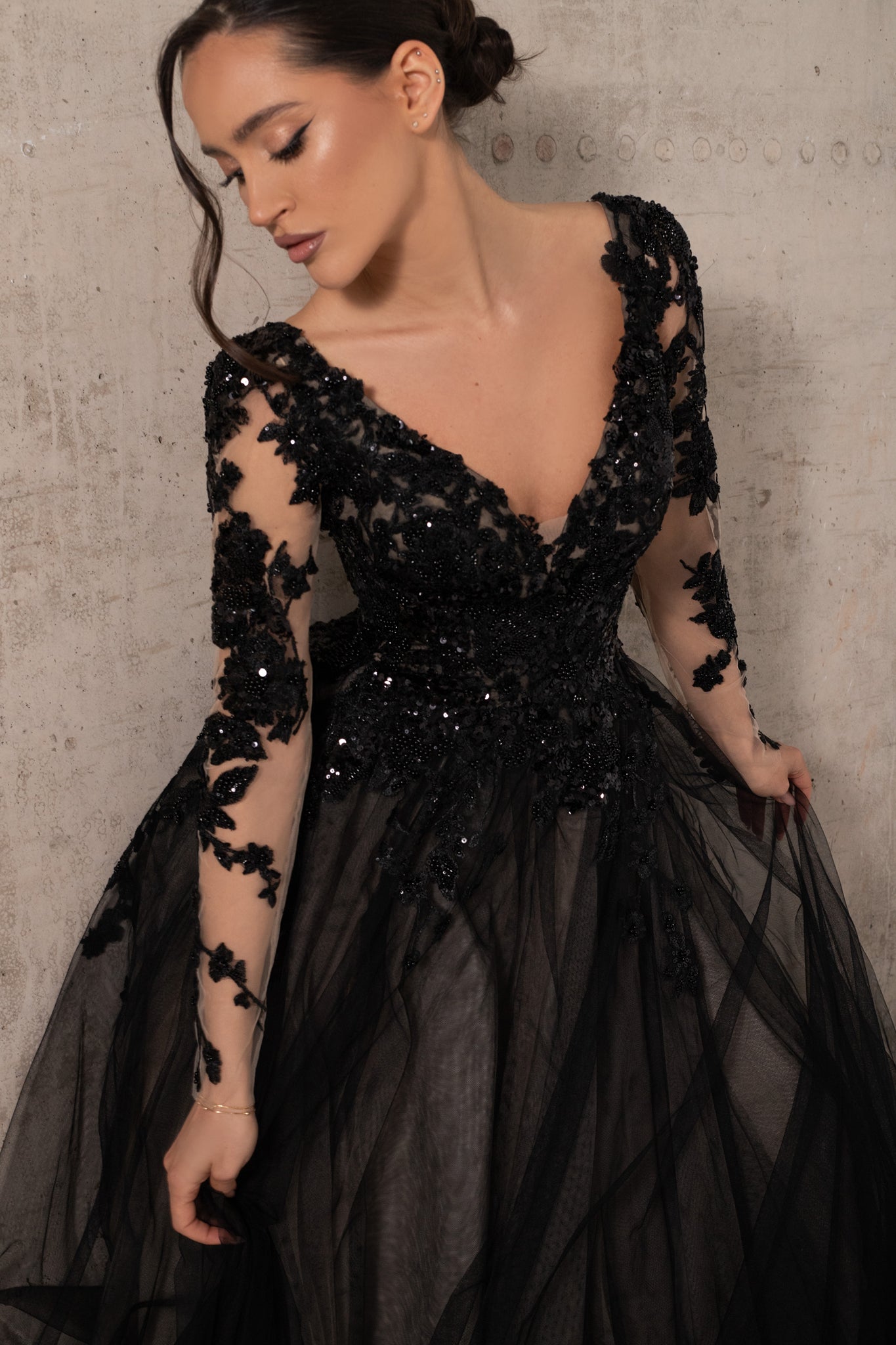 A woman wearing a black formal gown with sheer long sleeves, floral lace applique, a deep V-neckline, and a voluminous tulle skirt, posed against a textured concrete wall.