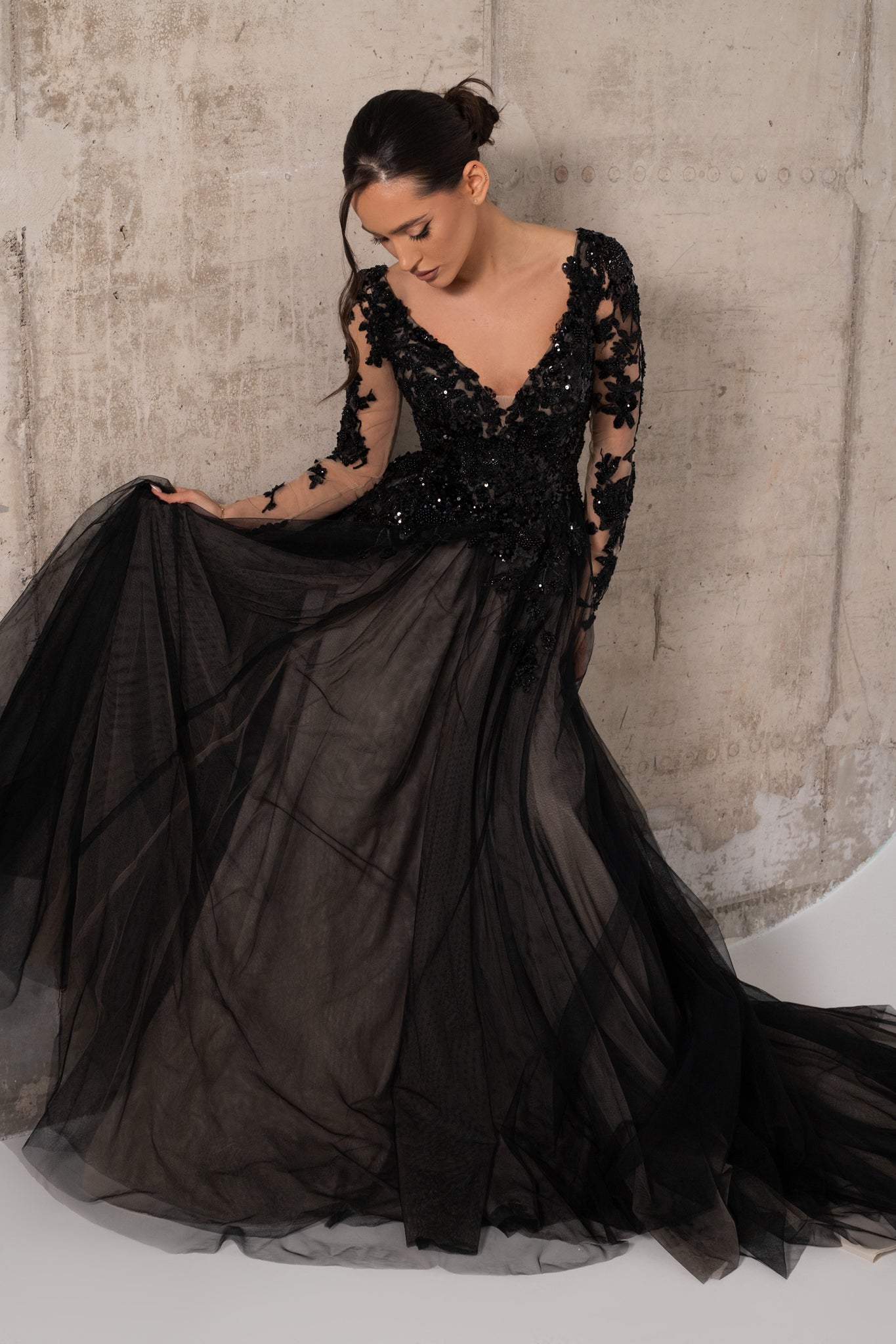 A woman wearing a dramatic black ball gown with sheer lace long sleeves, a deep V-neckline, and a voluminous tulle skirt, posed against a textured concrete wall.