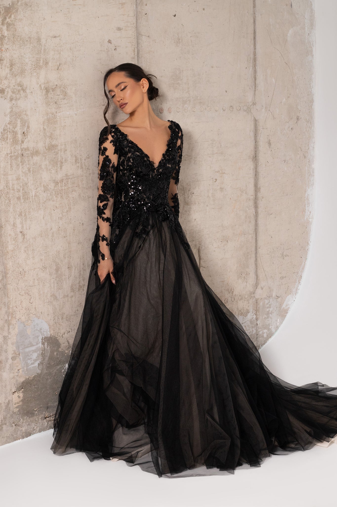 A woman models a black wedding ball gown with a deep V-neckline, heavily embellished beaded bodice, sheer lace sleeves, and a voluminous tulle skirt, posed against a textured concrete wall.