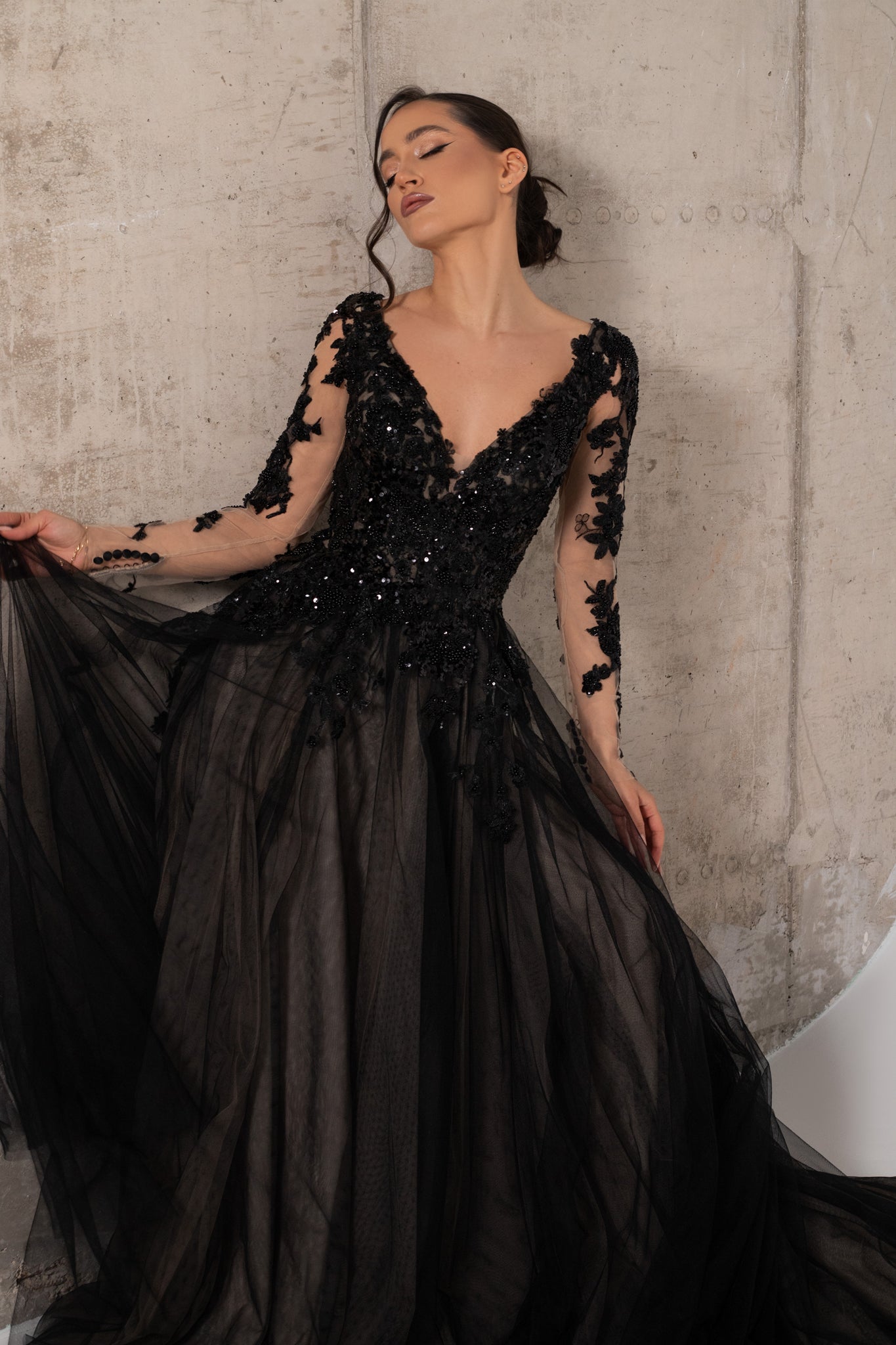 A woman modeling a black long sleeve wedding dress with intricate lace and sequined embellishments. The dress has deep plunging neckline and tulle skirt with multiple layers in champagne and black color.