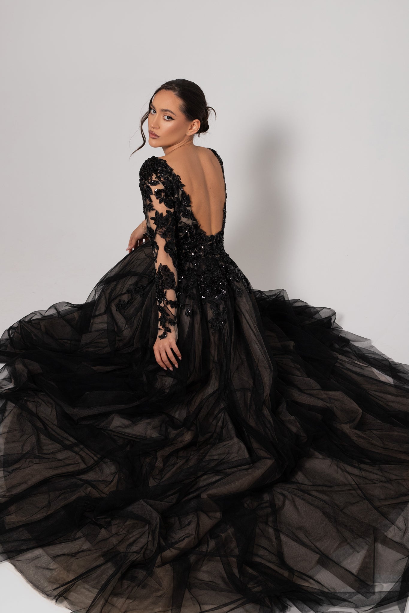 A model wearing a dramatic black ball gown with a tulle skirt, beaded long sleeves, and a deep open back, posed against a light gray studio background.