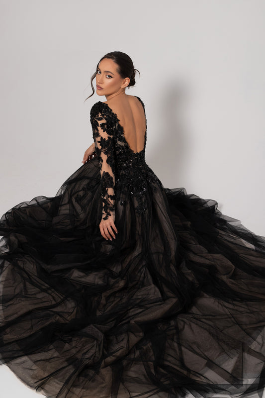 A model wearing a dramatic black ball gown with a tulle skirt, beaded long sleeves, and a deep open back, posed against a light gray studio background.