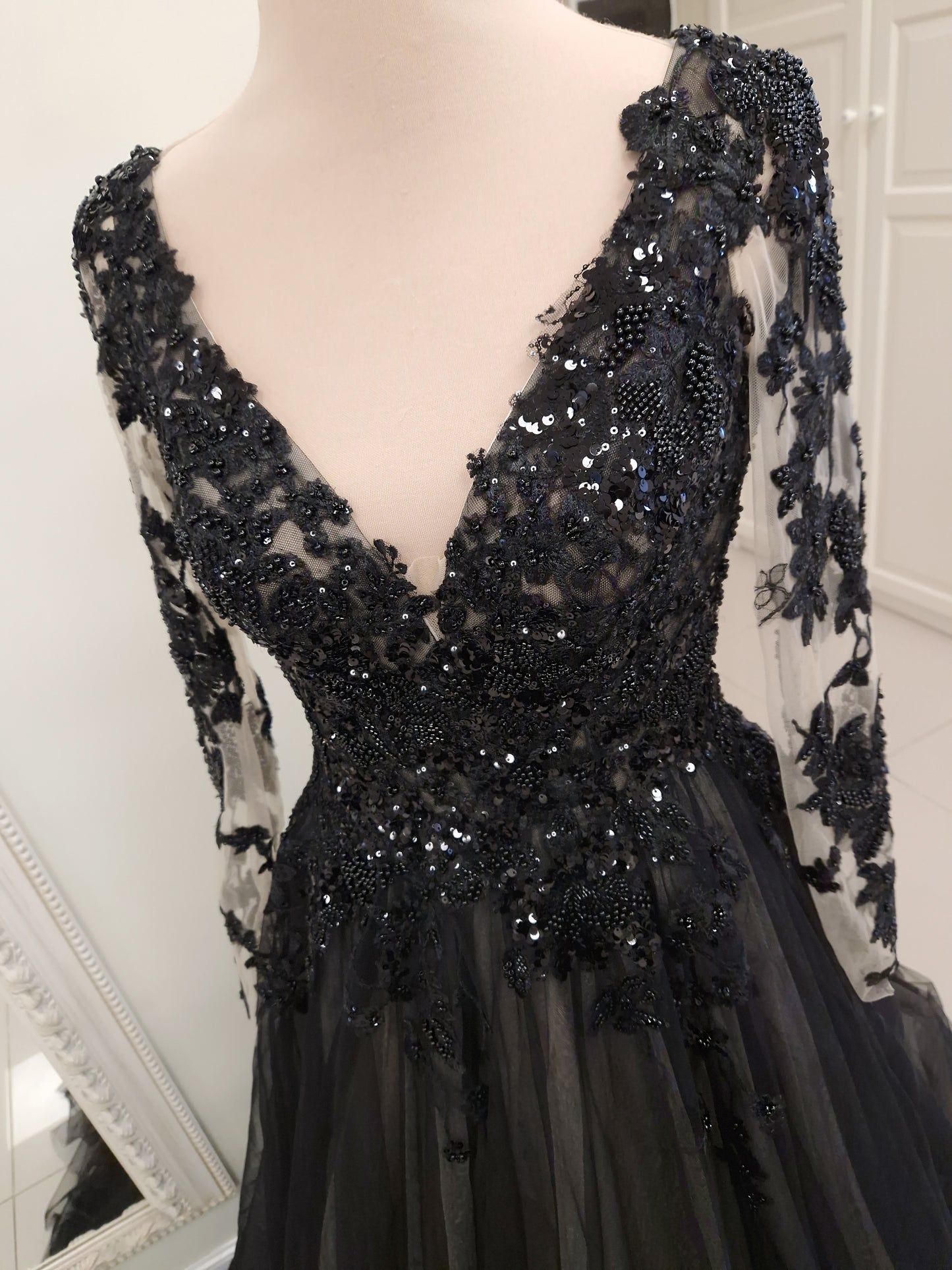 A black formal gown with long sheer sleeves, a deep V-neckline, and extensive black lace and bead embellishments, displayed on a mannequin.