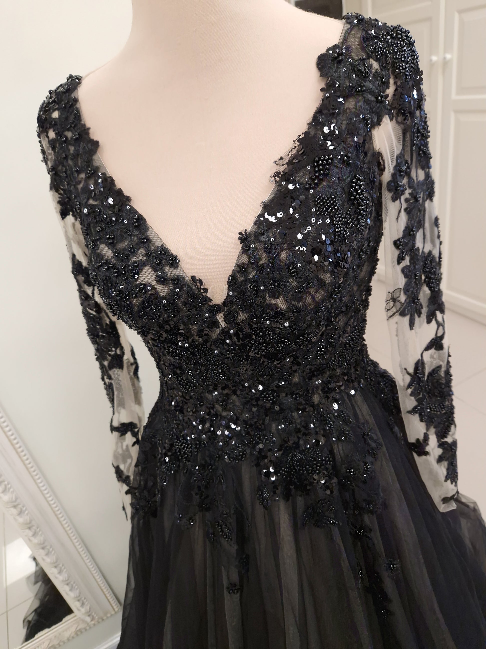 A black formal gown with long sheer sleeves, a deep V-neckline, and extensive black lace and bead embellishments, displayed on a mannequin.